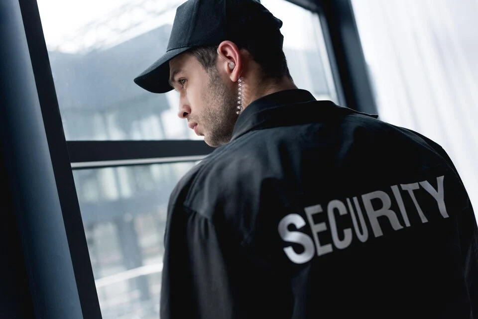 Security officer with earpiece and “SECURITY” jacket looking out a window